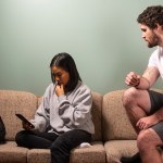 A man sits atop a couch's armrest as his romantic partner sits looking at her phone. The two are not facing one another and seem tense.