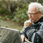 An older man with white hair is sat before his laptop, outside, showing distrust at what he sees.