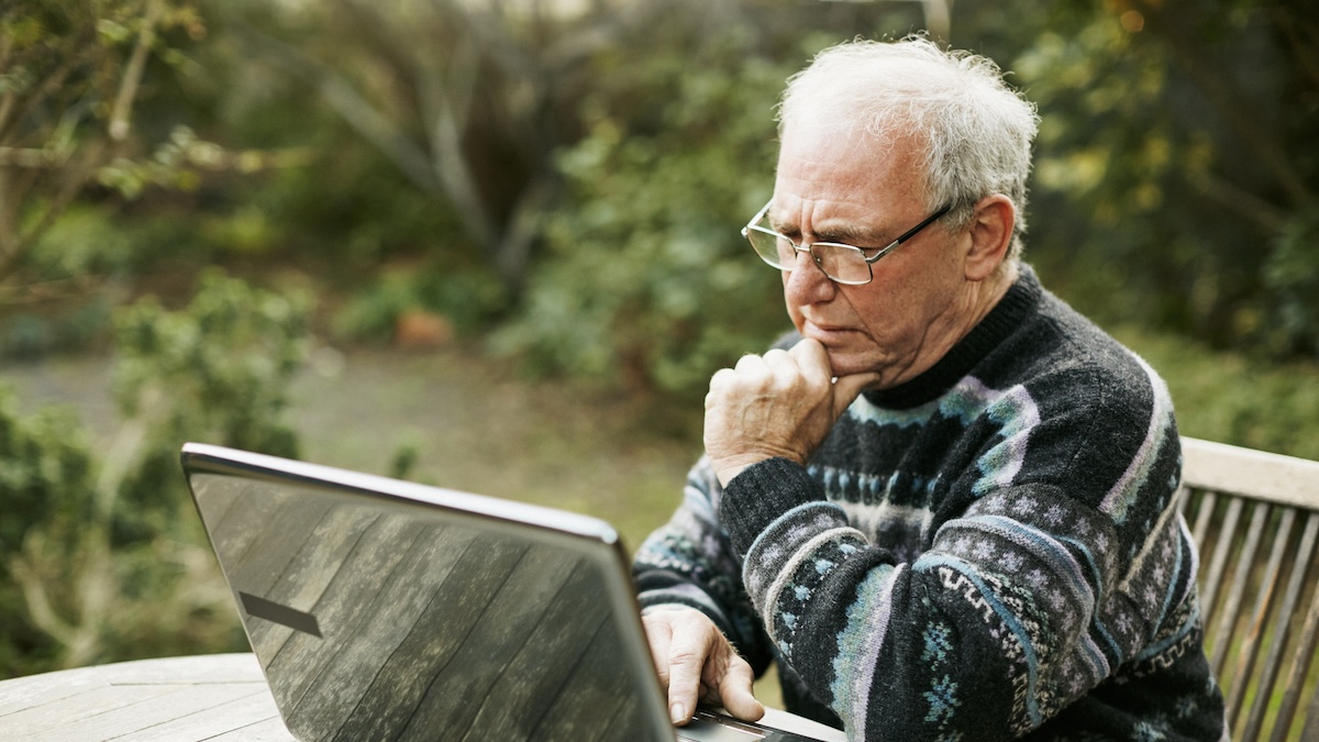 An older man with white hair is sat before his laptop, outside, showing distrust at what he sees.